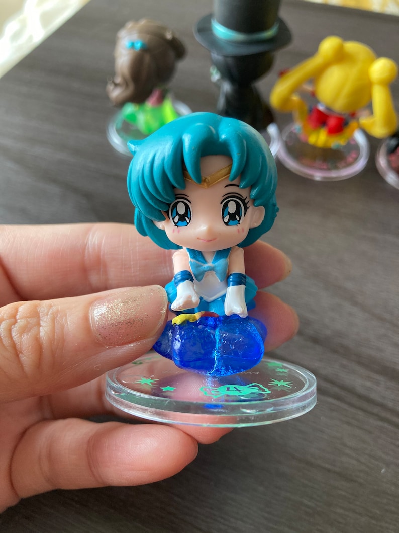 Puede incluir: Una peque&ntilde;a figura de un personaje con cabello azul y un atuendo azul, de pie sobre una base transparente. La figura tiene ojos grandes y forma parte de una colecci&oacute;n de figuras similares. Otras figuras son visibles en el fondo.