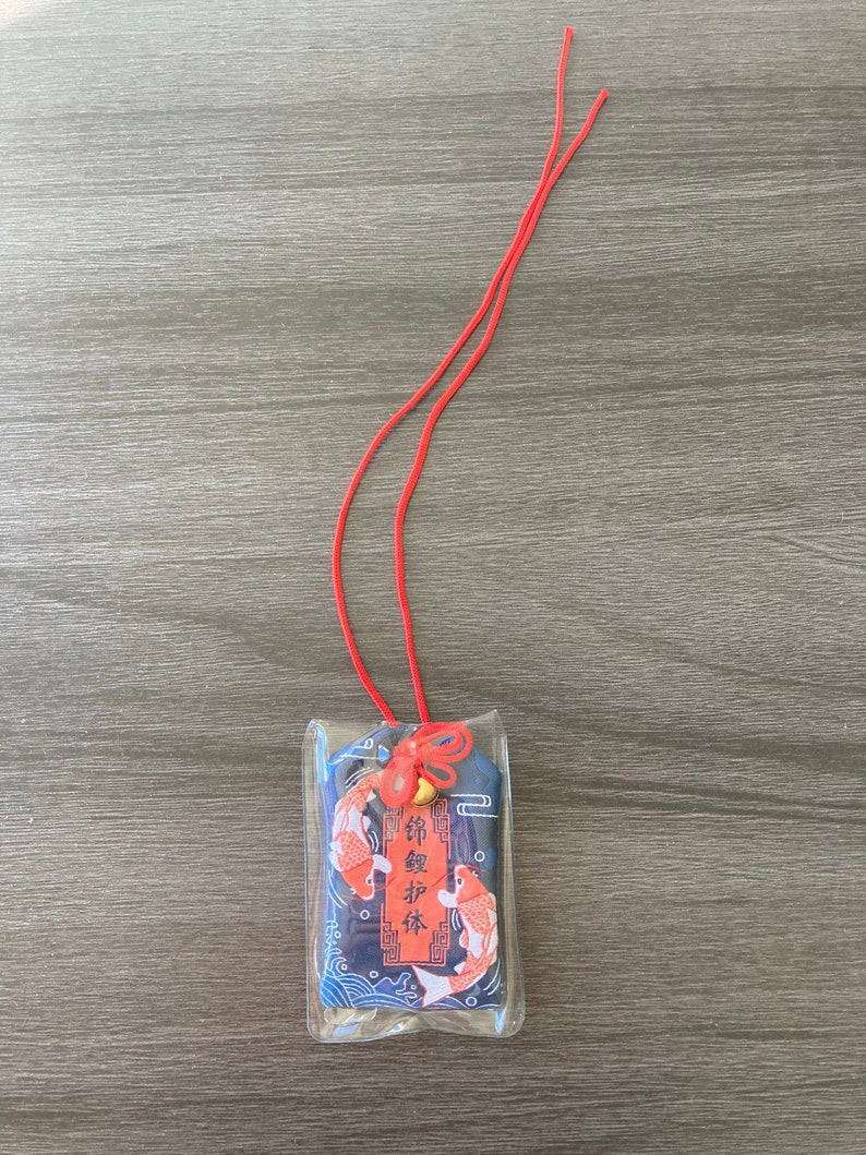 May include: A rectangular charm with a koi fish design, encased in clear plastic. The charm features two orange and white koi fish against a blue wave pattern. A red cord is attached at the top, along with a small bell and decorative knot.