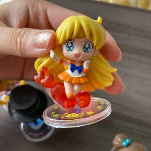 Puede incluir: Una peque&ntilde;a figura amarilla de un personaje con largo cabello rubio, ojos azules y un atuendo naranja. La figura est&aacute; de pie sobre una base circular transparente con un accesorio en forma de coraz&oacute;n. Otras figuras son visibles en el fondo.