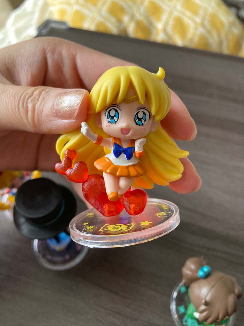 Puede incluir: Una peque&ntilde;a figura amarilla de un personaje con largo cabello rubio, ojos azules y un atuendo naranja. La figura est&aacute; de pie sobre una base circular transparente con un accesorio en forma de coraz&oacute;n. Otras figuras son visibles en el fondo.