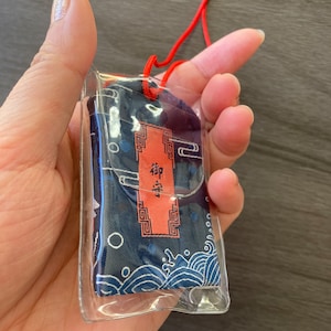 May include: A navy blue Japanese omamori charm with a red cord. The charm is in a clear plastic case. The fabric has a wave pattern and a red rectangular label with Japanese text.