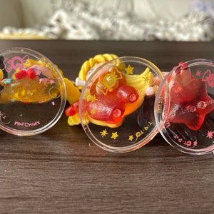 Puede incluir: Primer plano de tres colgantes circulares transparentes con dise&ntilde;os de dibujos animados coloridos. Los colgantes tienen detalles en rosa y amarillo, con un colgante rojo en forma de estrella a la derecha. El texto "MADE IN CHINA" es visible.