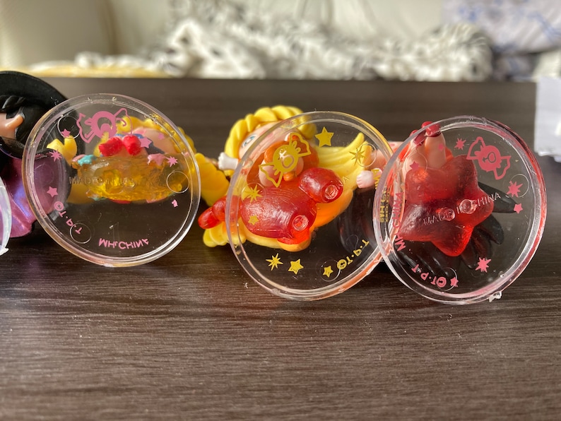 Puede incluir: Primer plano de tres colgantes circulares transparentes con dise&ntilde;os de dibujos animados coloridos. Los colgantes tienen detalles en rosa y amarillo, con un colgante rojo en forma de estrella a la derecha. El texto "MADE IN CHINA" es visible.