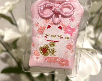 May include: Pink Japanese lucky charm, encased in clear plastic. The charm features a smiling cat design with floral accents and gold text. A pink cord is attached for hanging. The charm is a traditional symbol of good fortune.