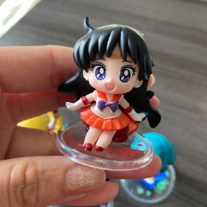 Puede incluir: Una peque&ntilde;a figura de un personaje con cabello negro, vistiendo un atuendo naranja y rojo con un lazo morado. La figura est&aacute; sobre una base circular transparente con detalles rojos. La figura tiene ojos grandes y expresivos.