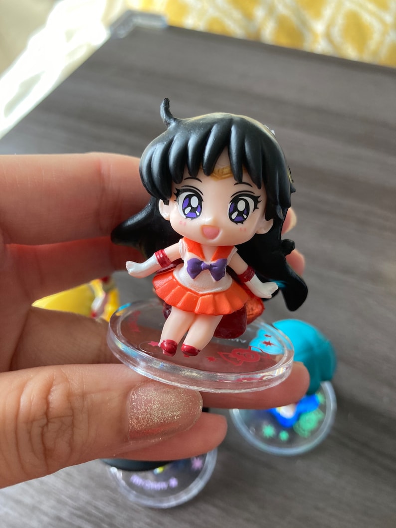Puede incluir: Una peque&ntilde;a figura de un personaje con cabello negro, vistiendo un atuendo naranja y rojo con un lazo morado. La figura est&aacute; sobre una base circular transparente con detalles rojos. La figura tiene ojos grandes y expresivos.