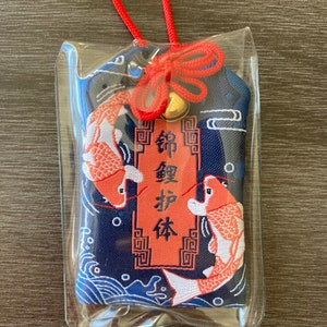 May include: A Japanese omamori charm in a clear plastic sleeve. The charm features two koi fish in orange and white, set against a navy blue background with wave patterns. The charm has a red cord and a small gold bell.
