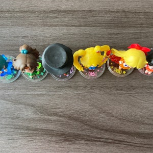 Puede incluir: Una colecci&oacute;n de seis peque&ntilde;as figuras, cada una representando un personaje con peinados y atuendos distintivos. Las figuras est&aacute;n dispuestas en fila sobre bases circulares transparentes. Los personajes tienen cabello azul, marr&oacute;n, amarillo y negro.