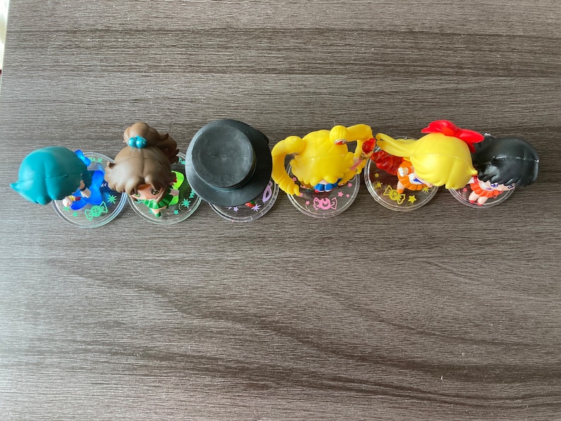 Puede incluir: Una colecci&oacute;n de seis peque&ntilde;as figuras, cada una representando un personaje con peinados y atuendos distintivos. Las figuras est&aacute;n dispuestas en fila sobre bases circulares transparentes. Los personajes tienen cabello azul, marr&oacute;n, amarillo y negro.