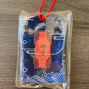 May include: A navy blue Japanese omamori charm in a clear plastic sleeve. The charm features a red rectangular tag with Japanese characters and decorative patterns. The design includes white waves and blue polka dots.