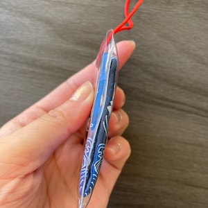 May include: A clear, plastic pendant with a blue and white patterned design, strung on a red cord. The pendant's design features a wave-like pattern. The cord is a bright red color.