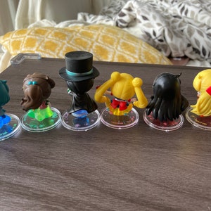 Puede incluir: Seis peque&ntilde;as figuras de personajes de anime, cada una con peinados y atuendos &uacute;nicos. Las figuras est&aacute;n sobre bases circulares transparentes con texto. Los personajes incluyen una figura con sombrero de copa y capa, y otros con pelo largo y lazos.