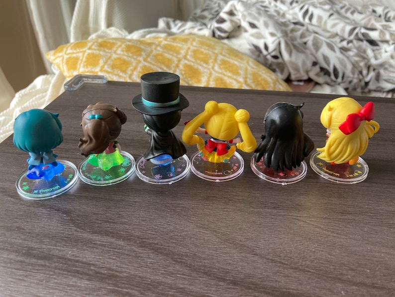 Puede incluir: Seis peque&ntilde;as figuras de personajes de anime, cada una con peinados y atuendos &uacute;nicos. Las figuras est&aacute;n sobre bases circulares transparentes con texto. Los personajes incluyen una figura con sombrero de copa y capa, y otros con pelo largo y lazos.