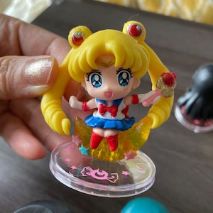 Puede incluir: Figura de Sailor Moon con cabello amarillo, falda azul y lazo rojo. La figura est&aacute; sobre una base transparente con texto rosa y un dise&ntilde;o de media luna. La figura sostiene un objeto dorado.