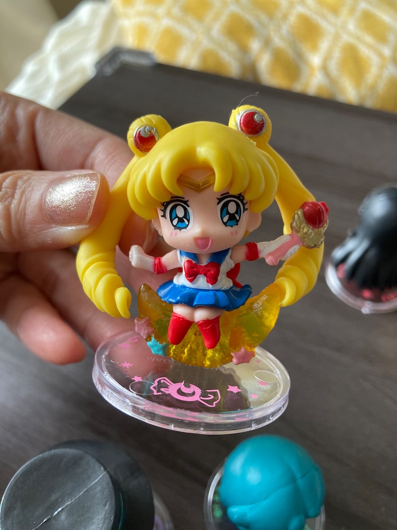 Puede incluir: Figura de Sailor Moon con cabello amarillo, falda azul y lazo rojo. La figura est&aacute; sobre una base transparente con texto rosa y un dise&ntilde;o de media luna. La figura sostiene un objeto dorado.