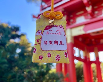 Könnte beinhalten: Gelber japanischer Glücksbringer mit einer lächelnden Katzenillustration und Text. Der Anhänger hat ein rotes Blumenmuster und eine kleine goldene Glocke. Er wird von einer gelben Schnur und einem Knoten gehalten.