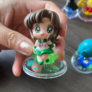 Puede incluir: Una peque&ntilde;a figura de un personaje con cabello casta&ntilde;o, ojos verdes y un atuendo verde y blanco. La figura est&aacute; sobre una base transparente con un brillo verde y detalles de estrellas. Otras figuras son visibles en el fondo.