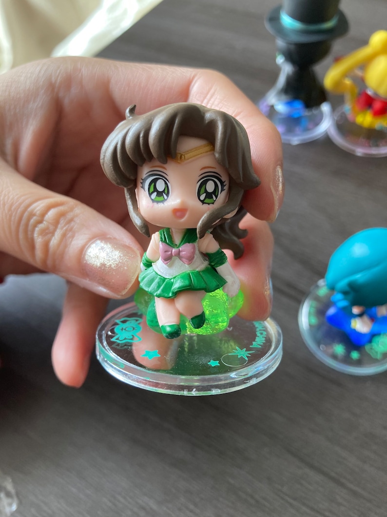Puede incluir: Una peque&ntilde;a figura de un personaje con cabello casta&ntilde;o, ojos verdes y un atuendo verde y blanco. La figura est&aacute; sobre una base transparente con un brillo verde y detalles de estrellas. Otras figuras son visibles en el fondo.
