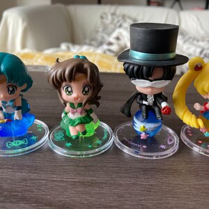 Puede incluir: Cuatro figuras de personajes de Sailor Moon. Cada figura est&aacute; sobre una base circular transparente con un dise&ntilde;o. Los personajes tienen ojos grandes y est&aacute;n en varias poses. Las figuras son en tonos de azul, verde, amarillo y negro.