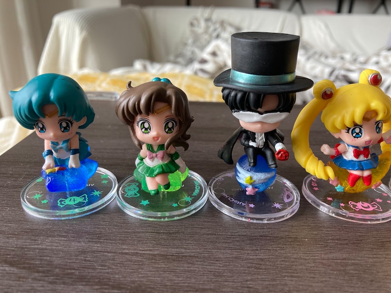 Puede incluir: Cuatro figuras de personajes de Sailor Moon. Cada figura est&aacute; sobre una base circular transparente con un dise&ntilde;o. Los personajes tienen ojos grandes y est&aacute;n en varias poses. Las figuras son en tonos de azul, verde, amarillo y negro.
