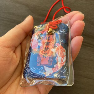 May include: A blue and red Japanese omamori charm with koi fish and text. The charm is in a clear plastic sleeve and has a red cord and a small gold bell.