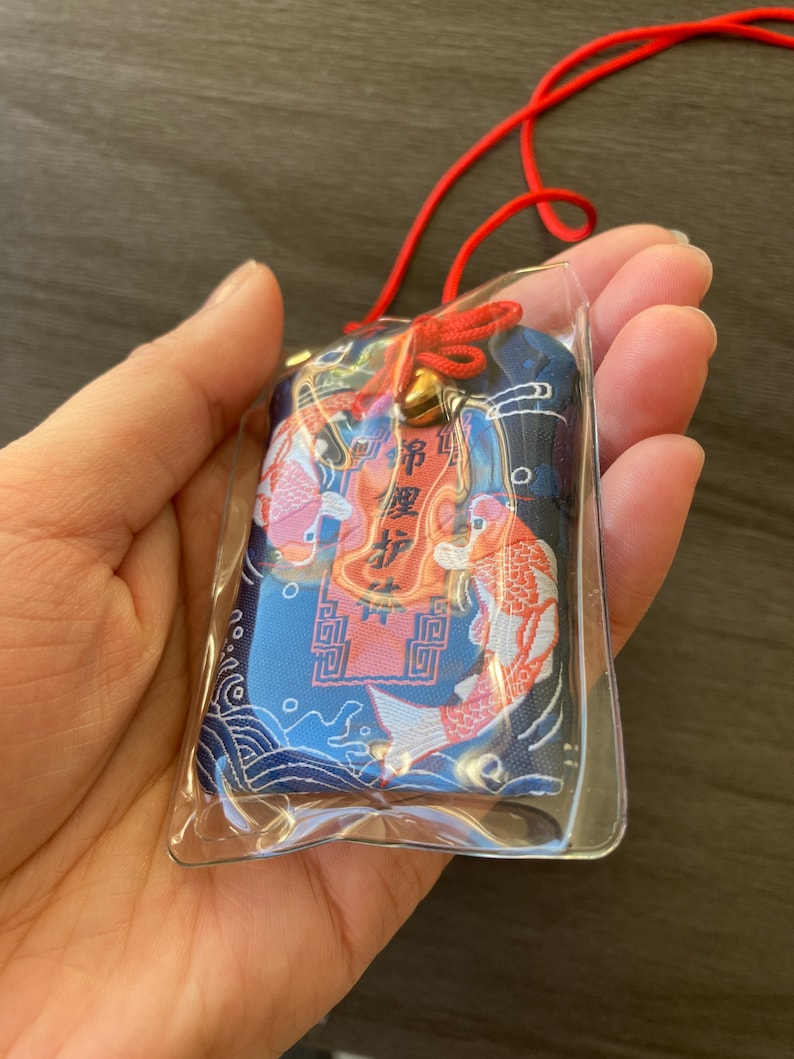 May include: A blue and red Japanese omamori charm with koi fish and text. The charm is in a clear plastic sleeve and has a red cord and a small gold bell.