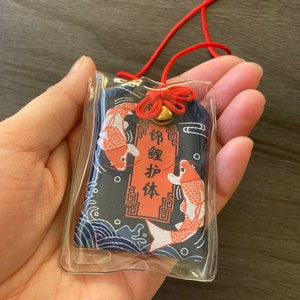 May include: A Japanese omamori charm in a clear plastic case. The charm features a navy blue background with koi fish and wave patterns. The text is in red and gold. A red cord and bell are attached.