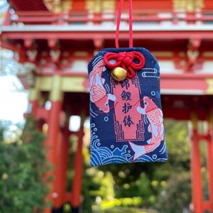May include: A navy blue Japanese omamori charm with a red cord and a small gold bell. The charm features two koi fish and Japanese text. The background shows a red torii gate.