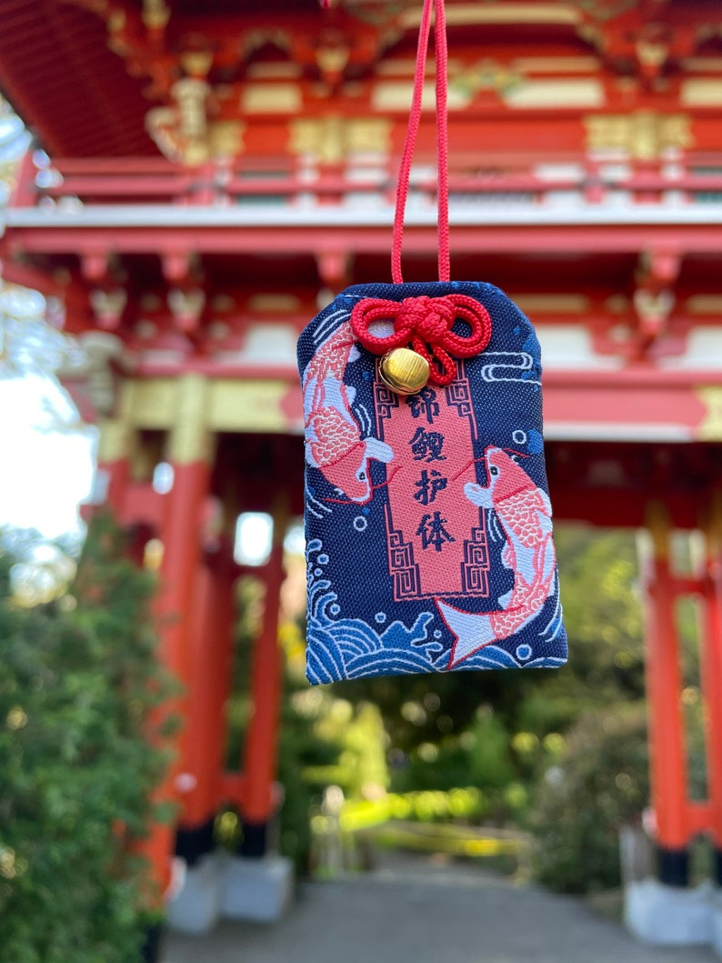 May include: A navy blue Japanese omamori charm with a red cord and a small gold bell. The charm features two koi fish and Japanese text. The background shows a red torii gate.