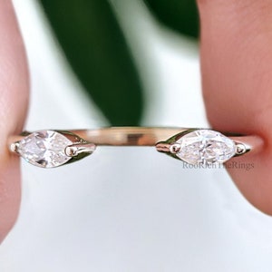 May include: A delicate gold ring with two marquise-shaped clear stones set on either side of the band. The ring is shown on a hand.