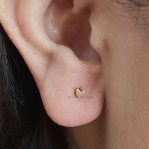May include: A close-up of an ear with a small, gold heart-shaped stud earring. The earring is positioned on the earlobe. The skin tone is light, and dark hair is visible in the background.
