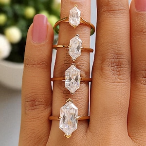 May include: Four gold rings with large, clear, geometric-cut gemstones are displayed on a hand. The rings are arranged vertically, each with a different size and shape of the gemstone. The hand has pink nail polish and is set against a soft background.