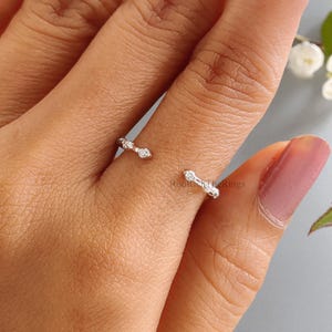 May include: Open-ended ring with a delicate design. The ring features small, sparkling gemstones set in a silver band. The ring is worn on a finger, showcasing its minimalist and elegant style. The gemstones add a touch of sparkle to the overall design.