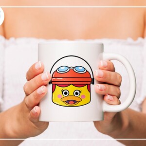 K&ouml;nnte beinhalten: Wei&szlig;e Keramik-Tasse mit einem Cartoon-Enten-Design. Die Ente hat ein gelbes Gesicht, einen orangefarbenen Hut und eine blaue Brille. Die Tasse wird von einer Person gehalten.