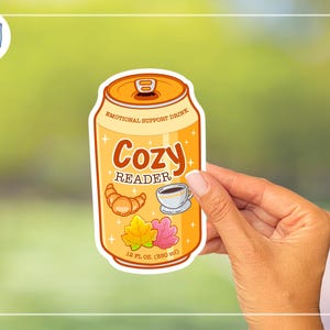 May include: A sticker featuring a cartoon can labeled "Emotional Support Drink." The can is orange with the words "Cozy Reader" and illustrations of a croissant, coffee cup, and leaves. It reads "12 fl oz (350 ml)."