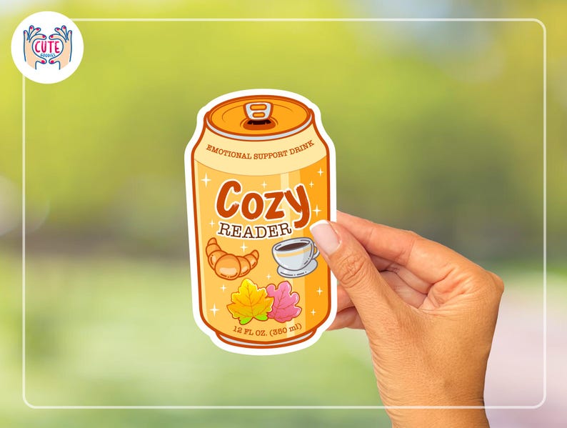 May include: A sticker featuring a cartoon can labeled "Emotional Support Drink." The can is orange with the words "Cozy Reader" and illustrations of a croissant, coffee cup, and leaves. It reads "12 fl oz (350 ml)."