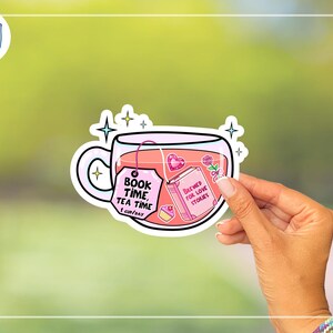 May include: A sticker featuring a teacup filled with pink tea. The teacup has a tag that reads "BOOK TIME, TEA TIME, 1 CUP/DAY." Inside the cup is a book that says "BREWED FOR LOVE STORIES." The sticker is held by a hand.