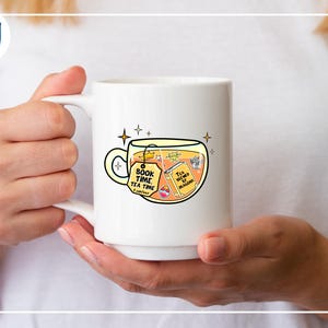 May include: White ceramic mug with a whimsical illustration of tea and books. The mug features the text "Book Time Tea Time" on a tag and "Tea brewed by magic" on a book. The design includes stars and a crown, suggesting a magical theme.