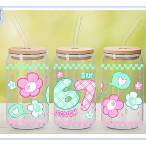 Puede incluir: Tres vasos de vidrio con tapas de madera y pajitas transparentes. Cada vaso presenta un dise&ntilde;o diferente: flores rosas y blancas, el n&uacute;mero "67" y un bocadillo con un coraz&oacute;n. Los vasos tienen un patr&oacute;n de cuadros rosas y verdes.