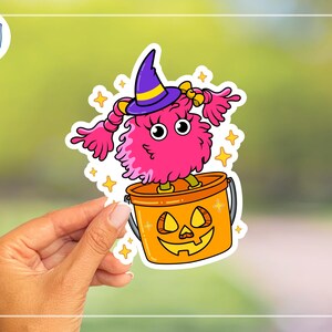 K&ouml;nnte beinhalten: Ein farbenfroher Halloween-Aufkleber mit einem rosa, flauschigen Monster, das einen lila Hexenhut mit gelbem Band tr&auml;gt. Das Monster steht in einem orangefarbenen K&uuml;rbis-Eimer. Gelbe Sterne umgeben das Monster und den Eimer.