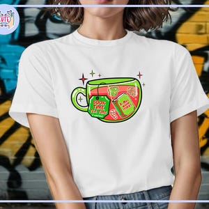 May include: White t-shirt featuring a whimsical illustration of a teacup filled with festive elements. The teacup design includes a tag that reads "Book Time Tea Time" and a book with the text "X-Mas Warm Stories".