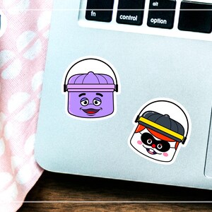 K&ouml;nnte beinhalten: Zwei Cartoon-Aufkleber auf einem Laptop. Einer ist ein lila Eimer mit einem l&auml;chelnden Gesicht. Der andere ist eine Figur mit schwarzem Hut, Maske und rotem Haar. Die Aufkleber sind wei&szlig; umrandet.