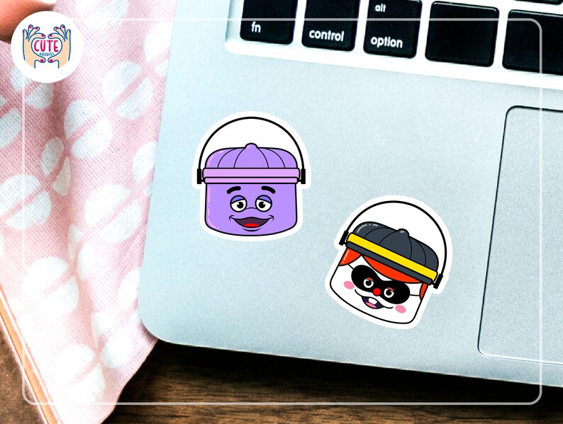 K&ouml;nnte beinhalten: Zwei Cartoon-Aufkleber auf einem Laptop. Einer ist ein lila Eimer mit einem l&auml;chelnden Gesicht. Der andere ist eine Figur mit schwarzem Hut, Maske und rotem Haar. Die Aufkleber sind wei&szlig; umrandet.