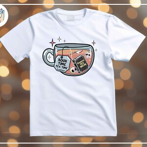 May include: White t-shirt featuring a whimsical design of a teacup filled with a book, knife, and skulls. The teacup has the text "Book Time, Tea Time" and a tag that says "I can't wait". A book in the teacup says "Haunted Tea Break".