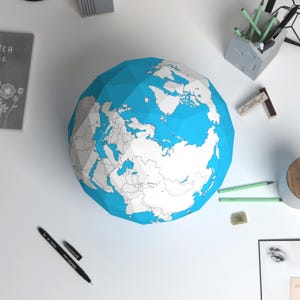 Puede incluir: Un globo terráqueo geométrico con océanos azules y continentes blancos se encuentra sobre una superficie blanca. Suministros de oficina, incluyendo bolígrafos, lápices y un pequeño portalápices de hormigón, rodean el globo. También se ve un bolígrafo negro y un cuaderno.