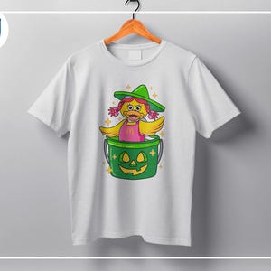 K&ouml;nnte beinhalten: Wei&szlig;es T-Shirt mit einer Cartoon-Ente, die einen gr&uuml;nen Hexenhut und ein rosa Kleid tr&auml;gt und in einem gr&uuml;nen K&uuml;rbis-Eimer sitzt. Das Design umfasst gelbe Fl&uuml;gel und Akzente, perfekt f&uuml;r Halloween. Das T-Shirt h&auml;ngt an einem Holzb&uuml;gel.