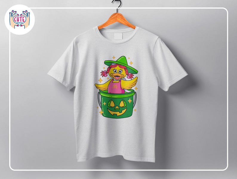 K&ouml;nnte beinhalten: Wei&szlig;es T-Shirt mit einer Cartoon-Ente, die einen gr&uuml;nen Hexenhut und ein rosa Kleid tr&auml;gt und in einem gr&uuml;nen K&uuml;rbis-Eimer sitzt. Das Design umfasst gelbe Fl&uuml;gel und Akzente, perfekt f&uuml;r Halloween. Das T-Shirt h&auml;ngt an einem Holzb&uuml;gel.