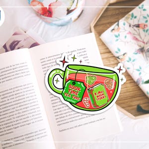 May include: A sticker featuring a green teacup with the text "Book Time Tea Time 1 Cup/Day" and "X-Mas Warm Stories" on a tag and a book. The sticker is placed on an open book with a floral design in the background.