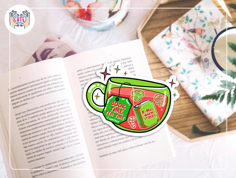 May include: A sticker featuring a green teacup with the text "Book Time Tea Time 1 Cup/Day" and "X-Mas Warm Stories" on a tag and a book. The sticker is placed on an open book with a floral design in the background.