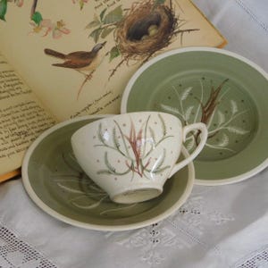 May include: A vintage tea set with a cream-colored teacup and saucer, and two green plates. The teacup and saucer feature a botanical design with red and green accents. An open book with a bird illustration is in the background.